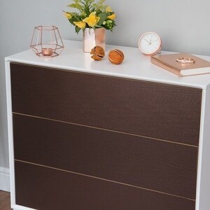 May include: A white chest of drawers with three drawers. The drawers are made of a dark brown material with a textured finish. The chest of drawers has a white top with a few decorative items on it, including a candle holder, a small clock, and a few decorative balls.