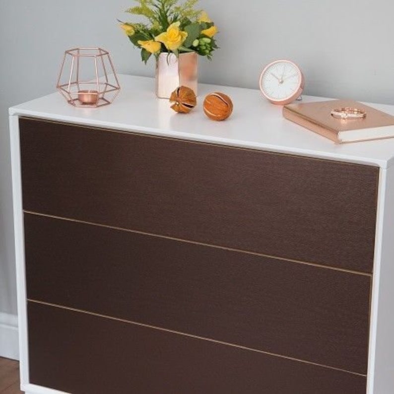 May include: A white chest of drawers with three drawers. The drawers are made of a dark brown material with a textured finish. The chest of drawers has a white top with a few decorative items on it, including a candle holder, a small clock, and a few decorative balls.