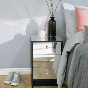 Pu&ograve; includere: Un comodino nero a specchio con tre cassetti. Il comodino ha una finitura in metallo martellato e pomelli di cristallo. Il comodino &egrave; accanto a un letto con un set di biancheria da letto bianco e grigio. Ci sono due vasi sul comodino, uno rosa e uno trasparente.