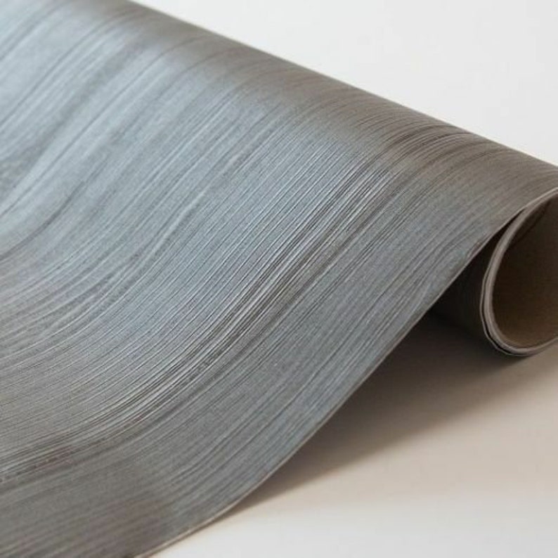 May include: A roll of gray wood-grain contact paper. The paper has a subtle sheen and a realistic wood grain pattern.