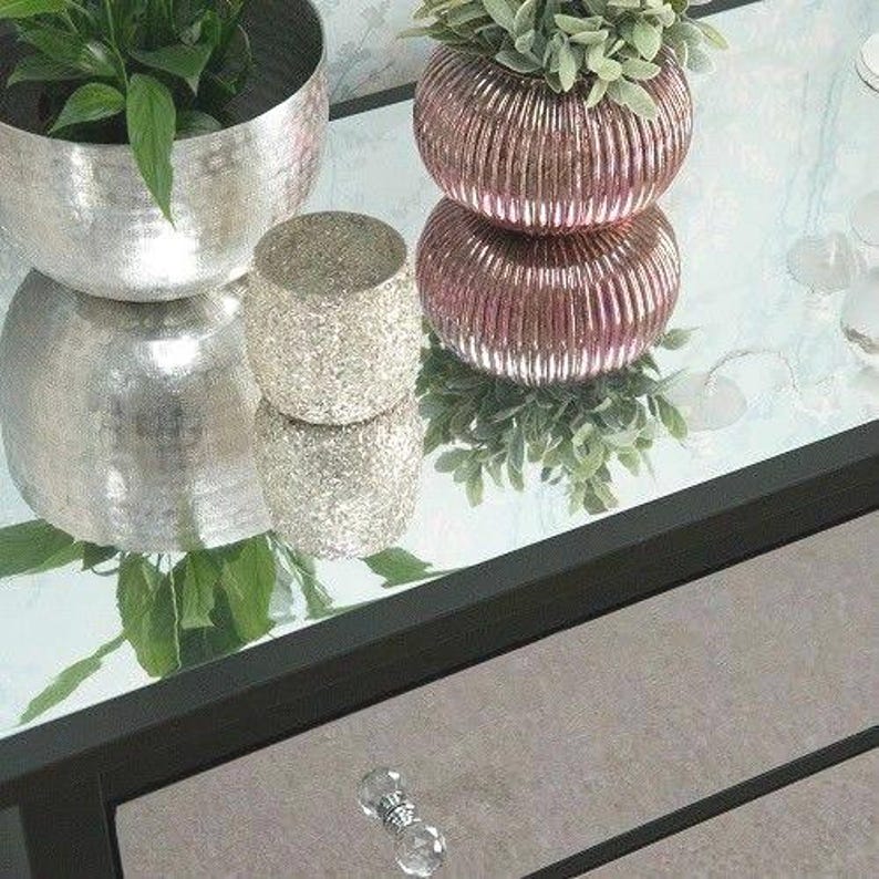 Pu&ograve; includere: Un tavolo nero con specchio con tre cassetti e vasi decorativi argentati e rosa con piante. Il tavolo ha una manopola argentata sul cassetto superiore.