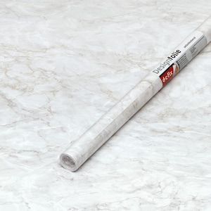 Pode incluir: Um rolo de filme autocolante Designfolie com padrão de mármore branco. O rolo está posicionado diagonalmente sobre uma superfície com um design de mármore correspondente. O rótulo do produto apresenta o texto "Designfolie" e "de fix".