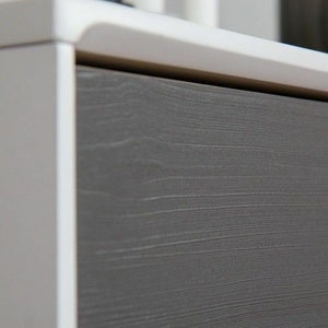 May include: Close-up of a drawer front with a wood-grain finish. The drawer is a light gray color with a white frame.
