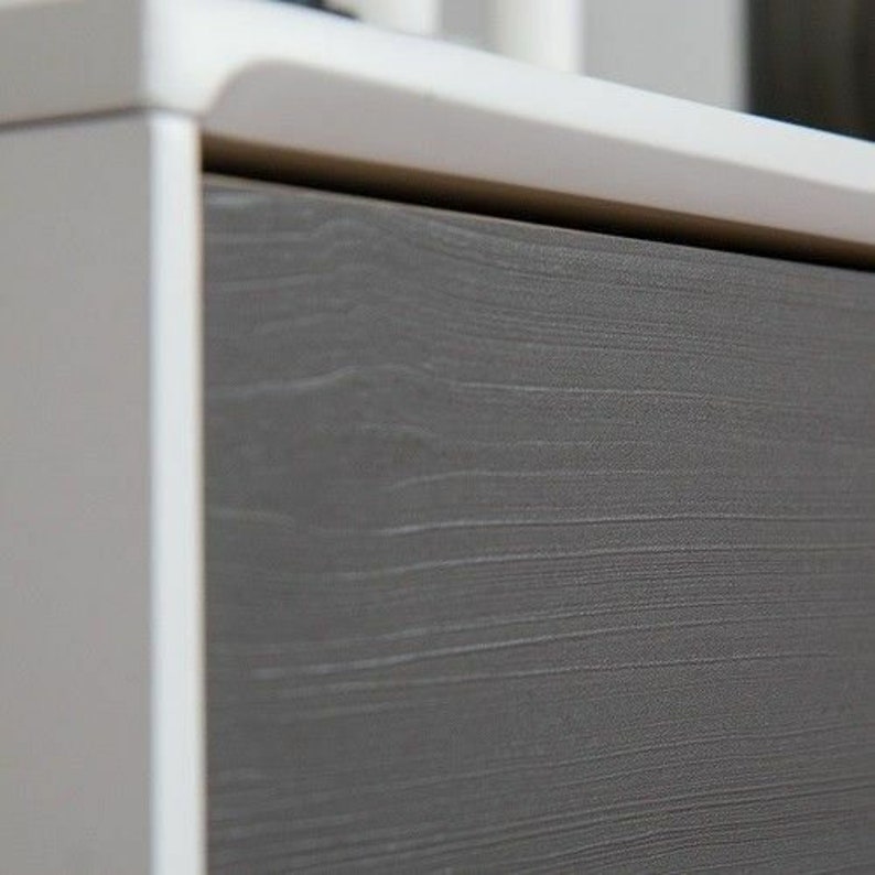 May include: Close-up of a drawer front with a wood-grain finish. The drawer is a light gray color with a white frame.