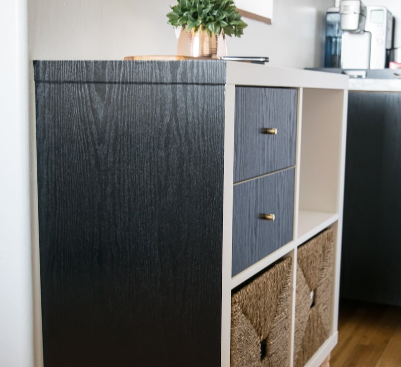 K&ouml;nnte beinhalten: Ein wei&szlig;es Regal mit schwarzem Holzmaserungsklebepapier auf der Vorder- und R&uuml;ckseite. Das Regal hat zwei Schubladen mit goldenen Kn&ouml;pfen und zwei offene Regalbretter mit geflochtenen K&ouml;rben.