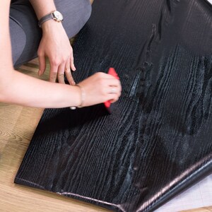 K&ouml;nnte beinhalten: Eine Person bringt schwarze Holzmaserung-Kontaktpapier mit einem roten Werkzeug auf eine Oberfl&auml;che an. Das Kontaktpapier hat eine abziehbare R&uuml;ckseite.