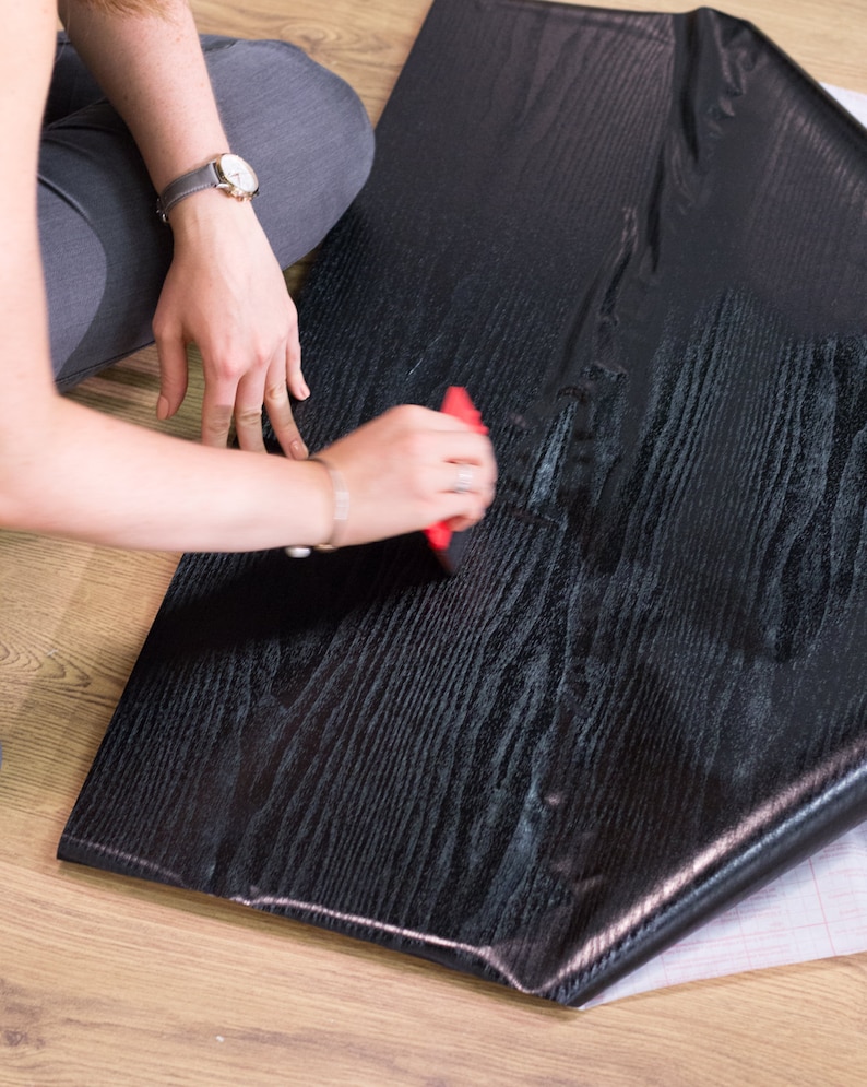 K&ouml;nnte beinhalten: Eine Person bringt schwarze Holzmaserung-Kontaktpapier mit einem roten Werkzeug auf eine Oberfl&auml;che an. Das Kontaktpapier hat eine abziehbare R&uuml;ckseite.