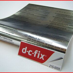 Może przedstawiać: Rolkę srebrnej folii odblaskowej z nadrukiem "CON FINITURA A SPECCHIO" i "FEUILLE REFLECTRICE". Folia jest oznaczona marką "d-c-fix&reg;" i numerem produktu "215-0005".