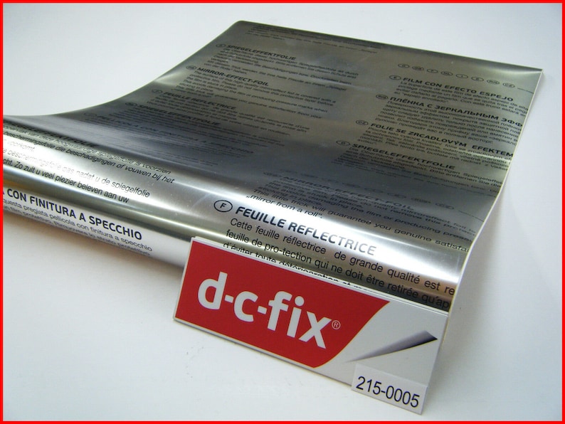 Może przedstawiać: Rolkę srebrnej folii odblaskowej z nadrukiem "CON FINITURA A SPECCHIO" i "FEUILLE REFLECTRICE". Folia jest oznaczona marką "d-c-fix&reg;" i numerem produktu "215-0005".
