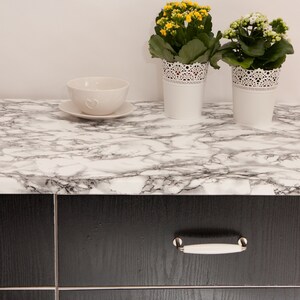 K&ouml;nnte beinhalten: Eine wei&szlig;e Keramiksch&uuml;ssel mit einem Herzdesign steht auf einer wei&szlig;en Marmor-Arbeitsplatte mit zwei Topfpflanzen. Die Pflanzen haben gr&uuml;ne Bl&auml;tter und gelbe und wei&szlig;e Bl&uuml;ten. Die Arbeitsplatte befindet sich &uuml;ber schwarzen Schr&auml;nken mit wei&szlig;en Griffen.