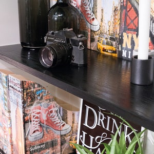 Pode incluir: Uma prateleira preta com uma câmera vintage, duas garrafas de vidro, uma vela branca e um castiçal preto. A prateleira é decorada com dois livros com tema da cidade de Nova York, uma placa que diz "Drink Local" e uma planta verde.