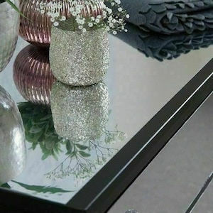 Pu&ograve; includere: Primo piano di una superficie a specchio con cornice nera. Due vasi decorativi argentati e rosa con fiori bianchi si riflettono nello specchio.