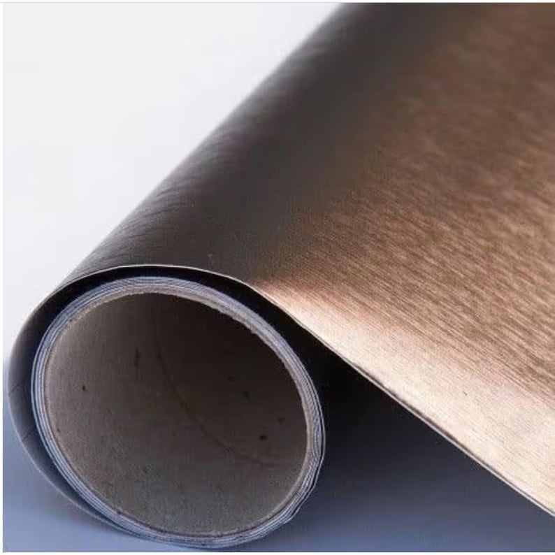 May include: A roll of brushed copper-coloured self-adhesive vinyl film. The film has a metallic finish and is designed for use on furniture, walls, and other surfaces.
