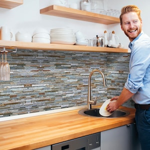 Puede incluir: Un hombre lava los platos en una cocina con un salpicadero de azulejos de piedra. El fregadero es de acero inoxidable con un grifo cromado. La encimera es de madera.