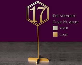 May include: Gold freestanding table number with a hexagonal design and the number 17. The table number is on a stand with a cross base. The image also shows the text "Freestanding Table Numbers" and the colors silver and gold.