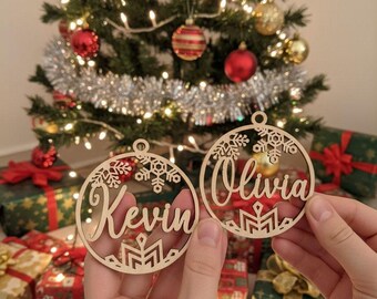 May include: Two wooden Christmas ornaments, one with the name "Kevin" and the other with "Olivia", are held in front of a decorated Christmas tree. The ornaments have snowflake and geometric designs. The tree is adorned with red and gold ornaments.