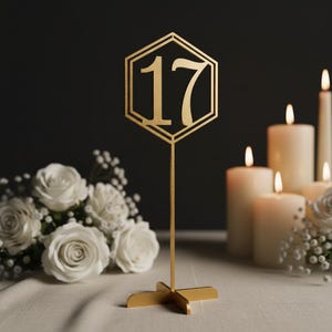 Op de afbeelding: Een gouden tafelnummer 17 in een zeshoekige lijst, met een standaard. Het nummer wordt weergegeven in een formeel schrift. De achtergrond omvat brandende kaarsen en witte rozen, wat duidt op een bruiloft of formeel evenement.
