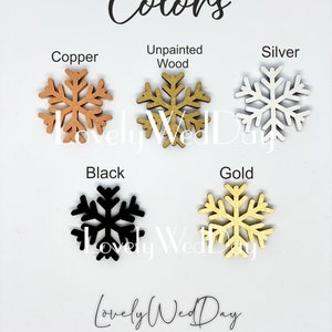 May include: Five wooden snowflake ornaments in different colors: copper, unpainted wood, silver, black, and gold. The snowflakes are arranged in two rows, with the copper, unpainted wood, and silver snowflakes in the top row and the black and gold snowflakes in the bottom row. The text "Colors" is written at the top of the image, and the text "Lovely Wed Day" is written at the bottom of the image.