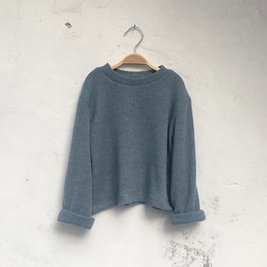 Puede incluir: Un suéter de punto acanalado azul grisáceo con cuello redondo y mangas largas. Las mangas están dobladas en las muñecas. El suéter está colgado de una percha de madera contra una pared blanca.