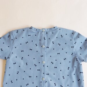 K&ouml;nnte beinhalten: Hellblaues kurz&auml;rmeliges Kleidungsst&uuml;ck mit einem Muster aus kleinen marineblauen Schleifen und Dreiecken. Die R&uuml;ckseite hat vier hellblaue Kn&ouml;pfe. Der Stoff ist wahrscheinlich Baumwolle. Kinderkleidung.
