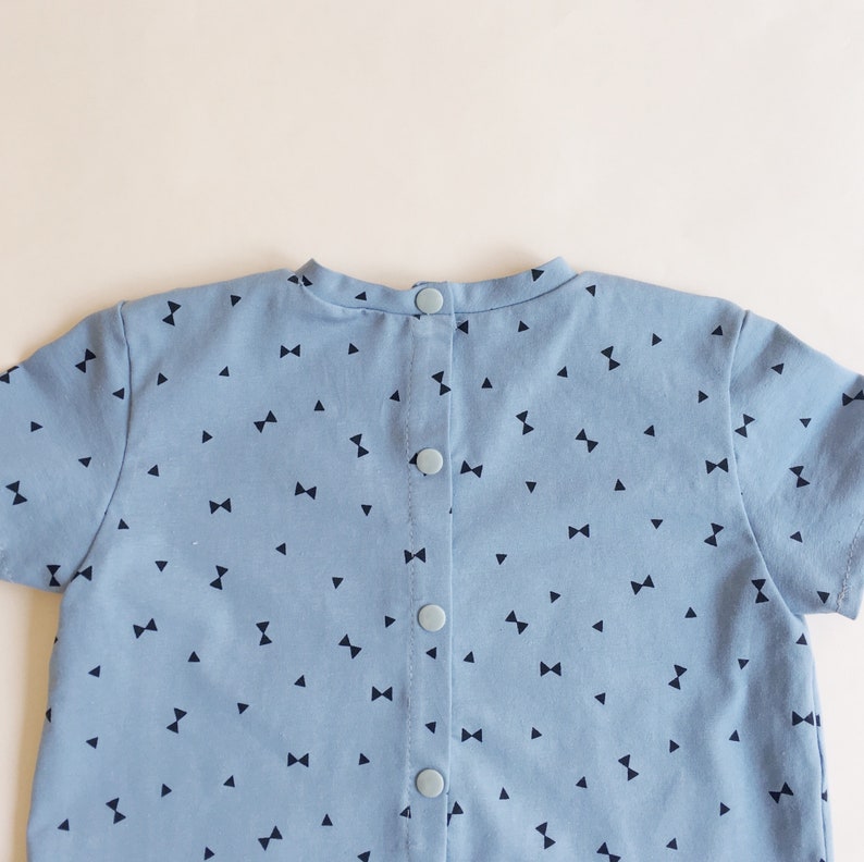 K&ouml;nnte beinhalten: Hellblaues kurz&auml;rmeliges Kleidungsst&uuml;ck mit einem Muster aus kleinen marineblauen Schleifen und Dreiecken. Die R&uuml;ckseite hat vier hellblaue Kn&ouml;pfe. Der Stoff ist wahrscheinlich Baumwolle. Kinderkleidung.