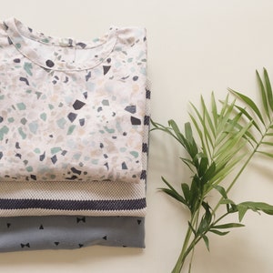K&ouml;nnte beinhalten: Drei gefaltete Hemden in einem Stapel. Das oberste Hemd hat einen wei&szlig;en Hintergrund mit einem Terrazzo-Muster in Blau-, Gr&uuml;n- und Schwarzt&ouml;nen. Das mittlere Hemd ist ein strukturiertes Strickteil mit gestreiftem Rand. Das untere Hemd ist grau mit schwarzen Schleifen- und Dreiecksmustern.