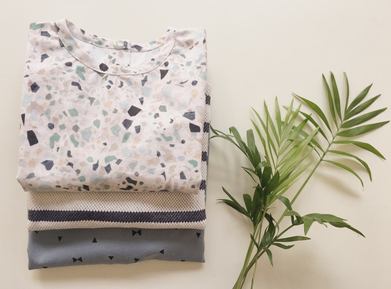K&ouml;nnte beinhalten: Drei gefaltete Hemden in einem Stapel. Das oberste Hemd hat einen wei&szlig;en Hintergrund mit einem Terrazzo-Muster in Blau-, Gr&uuml;n- und Schwarzt&ouml;nen. Das mittlere Hemd ist ein strukturiertes Strickteil mit gestreiftem Rand. Das untere Hemd ist grau mit schwarzen Schleifen- und Dreiecksmustern.