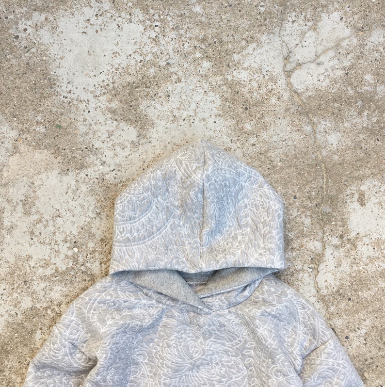Op de afbeelding: Een grijze hoodie met een wit paisley-patroon. De hoodie heeft een capuchon met trekkoord en een kangoeroezak.