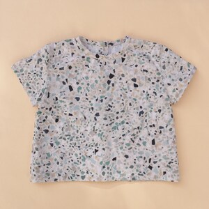 Könnte beinhalten: Ein kurzärmliges Oberteil mit Terrazzo-Muster in Beige-, Grün- und Schwarztönen. Das Rundhals-Shirt hat einen Knopfverschluss am Rücken und ist aus leichtem Stoff gefertigt. Das Oberteil ist ein lässiger Stil.