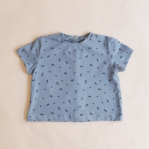 May include: A light blue short-sleeved shirt with a pattern of small, dark blue bow ties. The shirt has a round neckline and a button closure at the back of the neck. The fabric appears to be soft and lightweight, suitable for children's wear.
