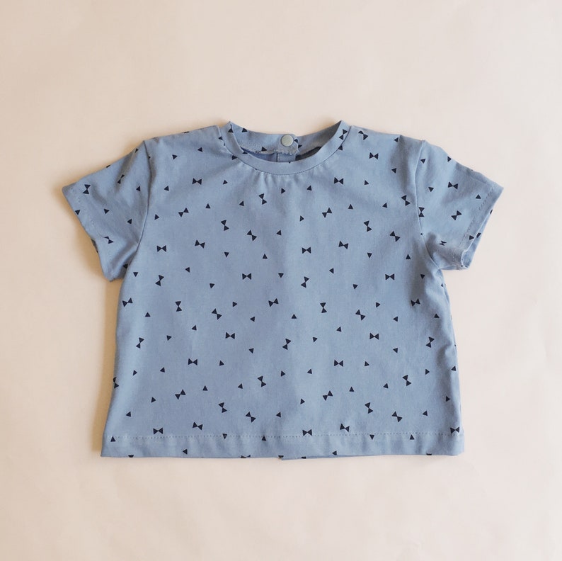K&ouml;nnte beinhalten: Hellblaues Kurzarm-T-Shirt mit einem Muster aus kleinen, dunkelblauen Schleifen. Das Shirt hat einen runden Ausschnitt und einen Knopfverschluss im Nacken. Der Stoff wirkt weich und leicht, geeignet f&uuml;r Kinderkleidung.