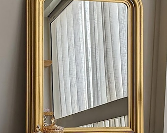 Puede incluir: Un espejo de pared rectangular con un marco dorado grueso y redondeado de acabado liso. El espejo tiene esquinas superiores redondeadas y un borde inferior plano, reflejando una ventana con cortinas blancas y un espacio interior oscuro.