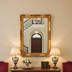 May include: A large gold-framed mirror reflects a hallway with a staircase and chandelier. A dark wood console table holds lamps, books, and flowers. Two red chairs sit either side of the table, creating a classic, elegant interior design.
