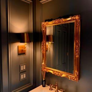 Pode incluir: Um espelho com moldura dourada está pendurado acima de uma pia branca com torneiras douradas. O espelho tem um design ornamentado. Uma pequena garrafa difusora preta está sobre a bancada. As paredes são cinza escuro com arandelas douradas.