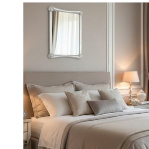 Puede incluir: Un espejo con marco plateado cuelga sobre una cama con cabecero beige. La cama está hecha con ropa de cama blanca y beige, incluyendo almohadas y una manta. Una lámpara con pantalla blanca está sobre una mesita de noche.