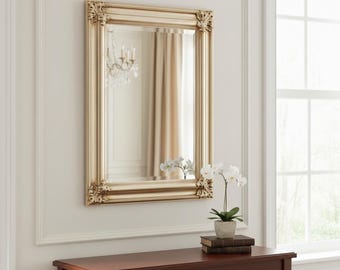 May include: A rectangular wall mirror featuring a metallic gold frame with ornate, carved detailing at each corner. The mirror has a beveled edge and is mounted on a plain, light-colored wall above a dark wooden console table with a potted plant.