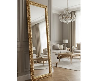May include: A tall, rectangular floor mirror featuring a thick, ornate gold frame with detailed carved scrollwork patterns. The mirror is positioned upright against a wall, reflecting the interior space of a room with light-colored furniture.
