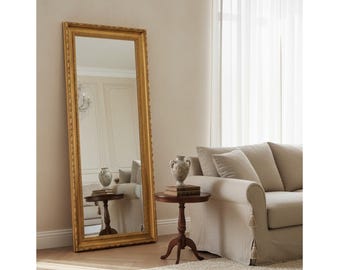 May include: A tall, rectangular floor mirror featuring a thick, ornate gold-toned frame with intricate detailing. The mirror is positioned upright against a wall, reflecting a small round wooden side table topped with a decorative ceramic vase.