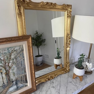 May include: A large, ornate gold mirror with a detailed frame is the focal point. A framed painting and two lamps with white shades flank the mirror. Small potted plants sit on a marble-topped table, adding a touch of greenery to the scene.