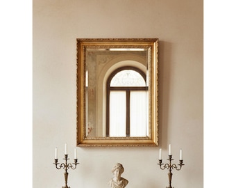 May include: A rectangular wall mirror with a decorative gold-toned frame featuring intricate carved detailing. The mirror reflects an arched window. Below the mirror are two ornate metal candelabras and a small stone-colored bust of a person.