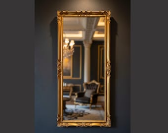 May include: A rectangular wall mirror featuring an ornate gold frame with intricate carved detailing along the edges. The reflective glass surface displays a clear view of a room interior, including a dark armchair, a pillar, and a chandelier.