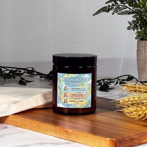 May include: A dark brown jar candle with a black lid, labeled "Psychological Sigh." The label features instructions for deep breathing. The candle sits on a wooden board with a white cloth and greenery in the background.