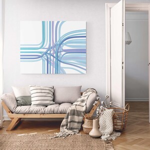 May include: A modern living room scene featuring a light wood futon sofa with gray cushions and throw pillows. A canvas art piece with blue and purple lines hangs above the sofa. A woven rug, baskets, and decorative items complete the space.