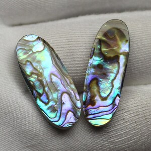 May include: Two oval-shaped abalone shells with iridescent colors of blue, green, purple, and gold. The shells have a smooth, polished surface and a natural, organic pattern. The shells are likely used for jewelry or decorative purposes.