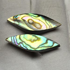 May include: Two iridescent abalone shell cabochons. The cabochons are teardrop-shaped with a dark border. The shells display a range of colors, including green, yellow, and blue, with swirling patterns. These are suitable for jewelry making.