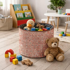 Op de afbeelding: Een ronde speelgoedbak met een patroon in rood, roze en grijs, gevuld met speelgoed, waaronder een pluchen eend, houten blokken en een teddybeer. Een houten speelgoedauto en een regenboogboog liggen op de vloer.