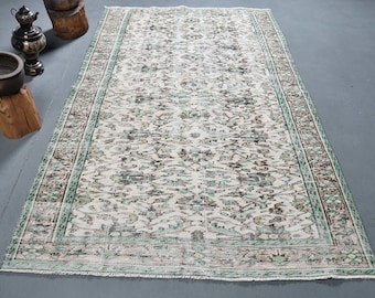Puede incluir: Una alfombra vintage con fondo blanco y un patrón floral en tonos verdes, marrones y rosas. La alfombra tiene un aspecto desgastado y un borde con flecos.