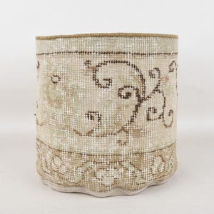 Op de afbeelding: Een cilindrische tapijtmand in beigetinten met een decoratief patroon. De mand heeft een herhalend bloemmotief in bruin en beige, een geschulpte basis en een bruine rand rond de bovenrand.