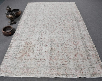 Puede incluir: Una alfombra vintage de color crema con un patrón floral en tonos rosa y gris. La alfombra tiene un borde con flecos y está tejida con un aspecto desgastado.