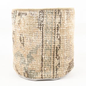 May include: A cylindrical, woven basket with a neutral color palette. The basket features a textured, patterned design with vertical stripes in shades of brown, beige, and gray. The base is a solid gray color.